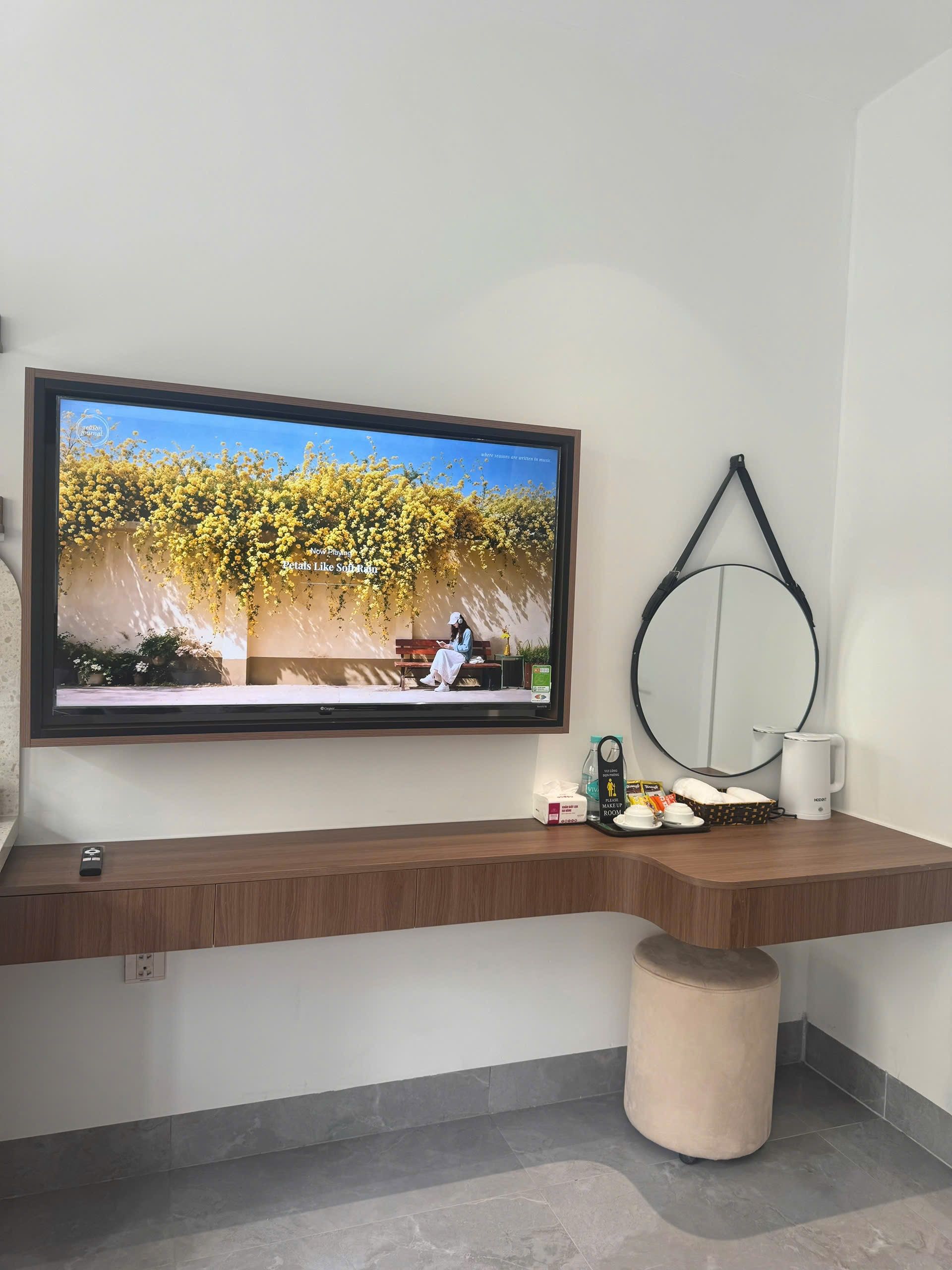 A modern bathroom with a large TV mounted on the wall, a round mirror, a wooden vanity, and a small stool.