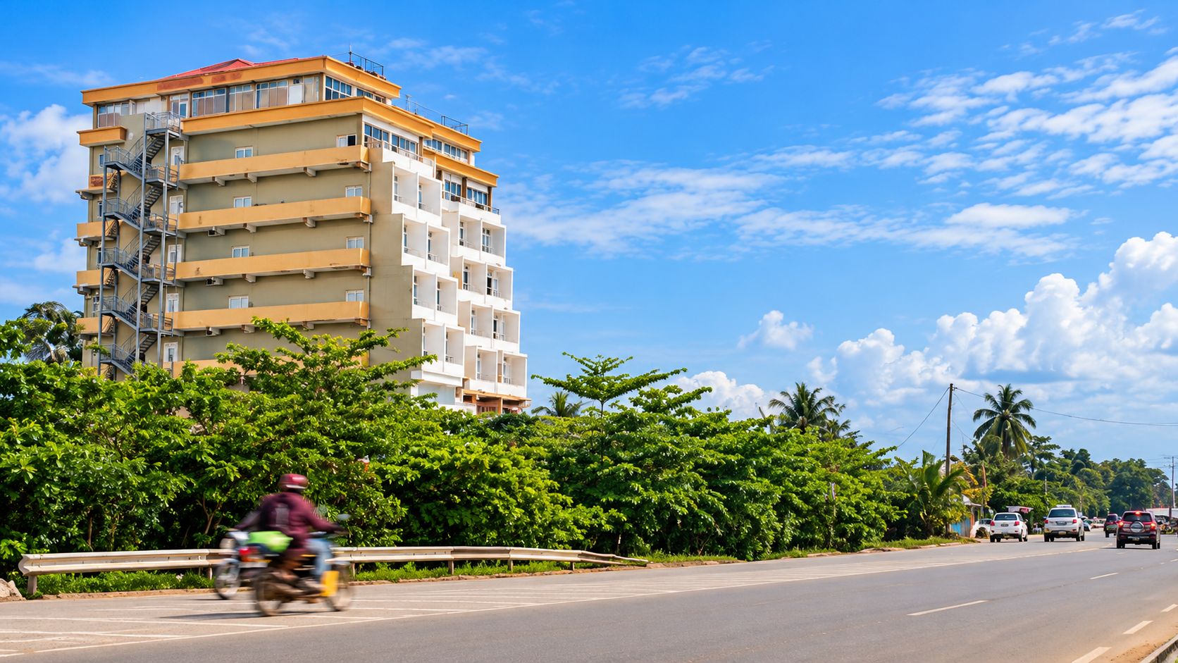 A multi-story building with balconies, a road with vehicles, and greenery.