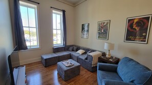 Sala de estar con sofá modular, otoman y sillón azul. Dos ventanas con cortinas, una lámpara en una mesita auxiliar y carteles enmarcados en la pared.