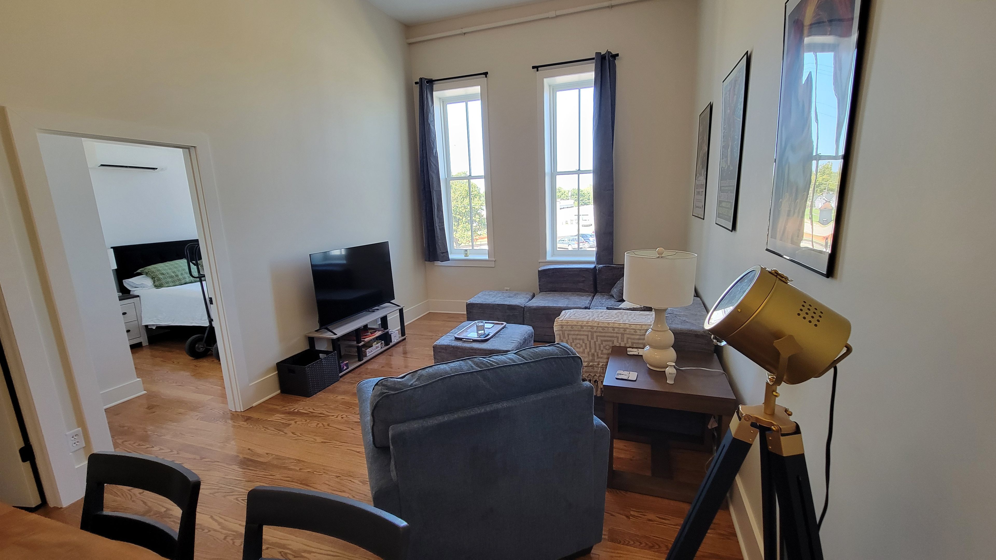 Un salon comprenant un téléviseur, un canapé, un pouf, une lampe et un bureau avec une lampe. Il y a des fenêtres avec des rideaux et une porte qui donne sur une autre pièce.