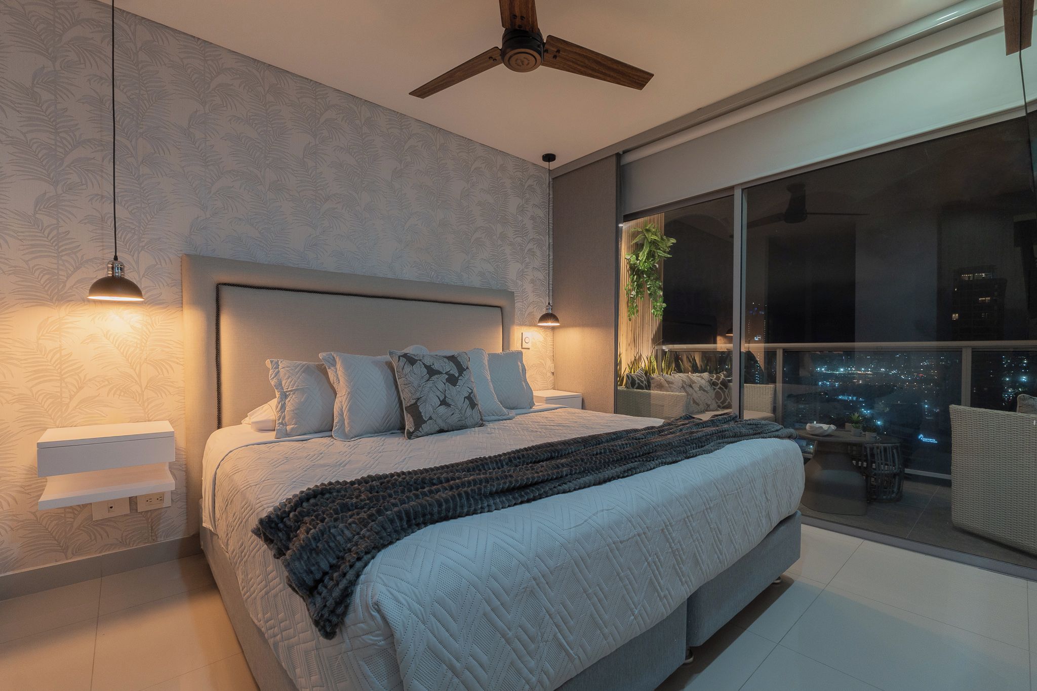 Ein modernes Schlafzimmer mit einem großen Bett, Nachttischlampen, einem Deckenventilator und Blick auf die Stadt durch ein großes Fenster.