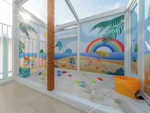 Una habitación con un mural con temática playera, una estructura de juego con cuerdas y un arenero con juguetes.