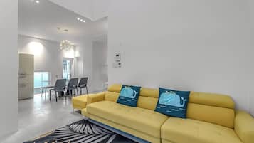 A modern living room with a yellow sofa, a patterned rug, and a dining area with chairs and a table.