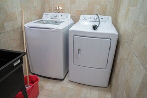 Two front-loading washing machines in a bathroom with tiled walls and a red trash bin. - Gulf Residence Apt1 (La Romain)