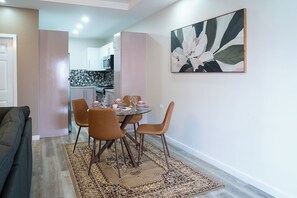 A dining area with a round table set for four, surrounded by four chairs, and a kitchen in the background. - Gulf Residence Apt1 (La Romain)