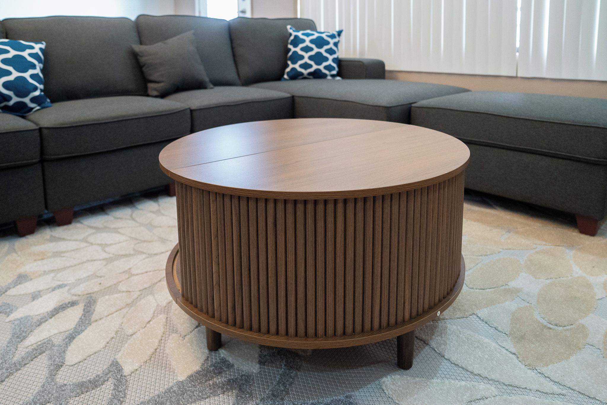 A round wooden coffee table with a ribbed design, placed on a patterned rug in a living room with a gray sectional sofa and patterned throw pillows.