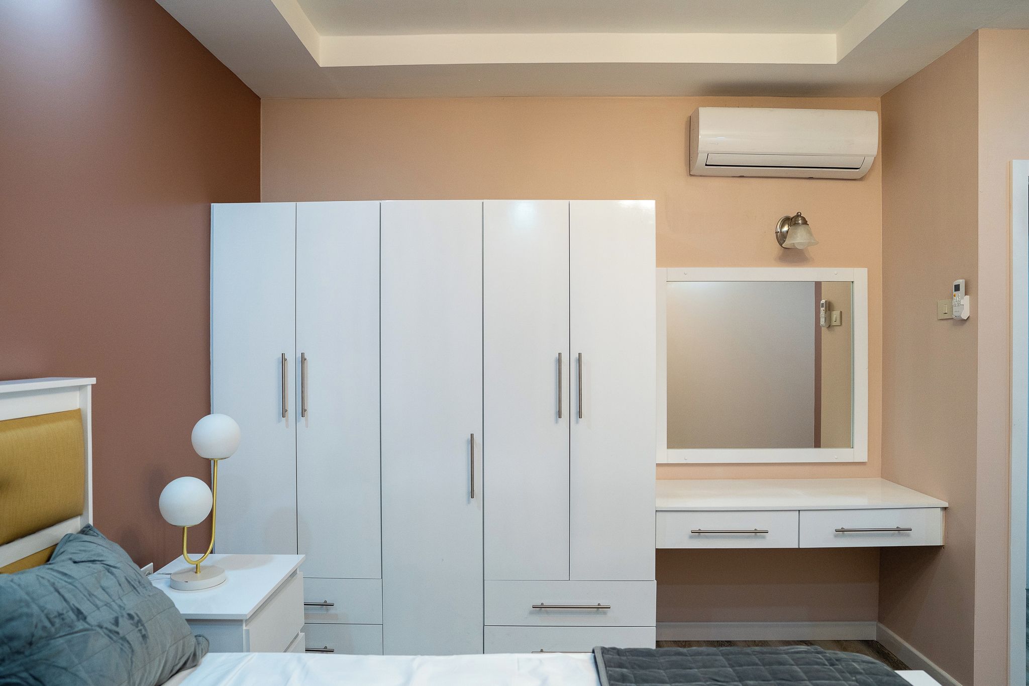 A bedroom with a bed, a wardrobe, a bedside table with a lamp, a mirror, and an air conditioning unit.
