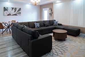 A modern living room with a sectional sofa, a round coffee table, and a dining area with a glass table and chairs. - Gulf Residence Apt1 (La Romain)