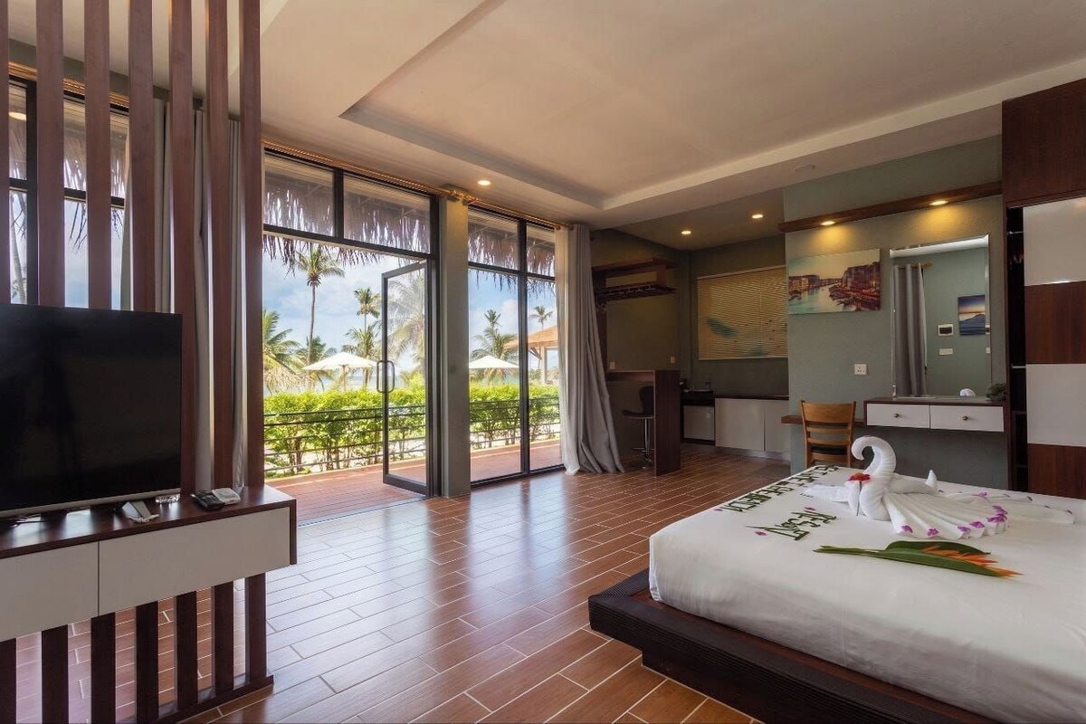 A hotel room with a large bed, a television, a desk, and a view of palm trees through the windows.