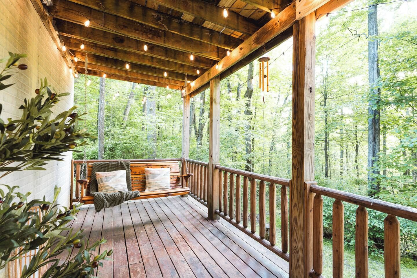 A wooden deck with a bench and hanging lights, surrounded by a forest.