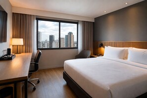 Ett hotellrum med en stor säng, ett skrivbord med en lampa, en stol, ett nattduksbord med en telefon och utsikt över stadsbilden genom fönstret.