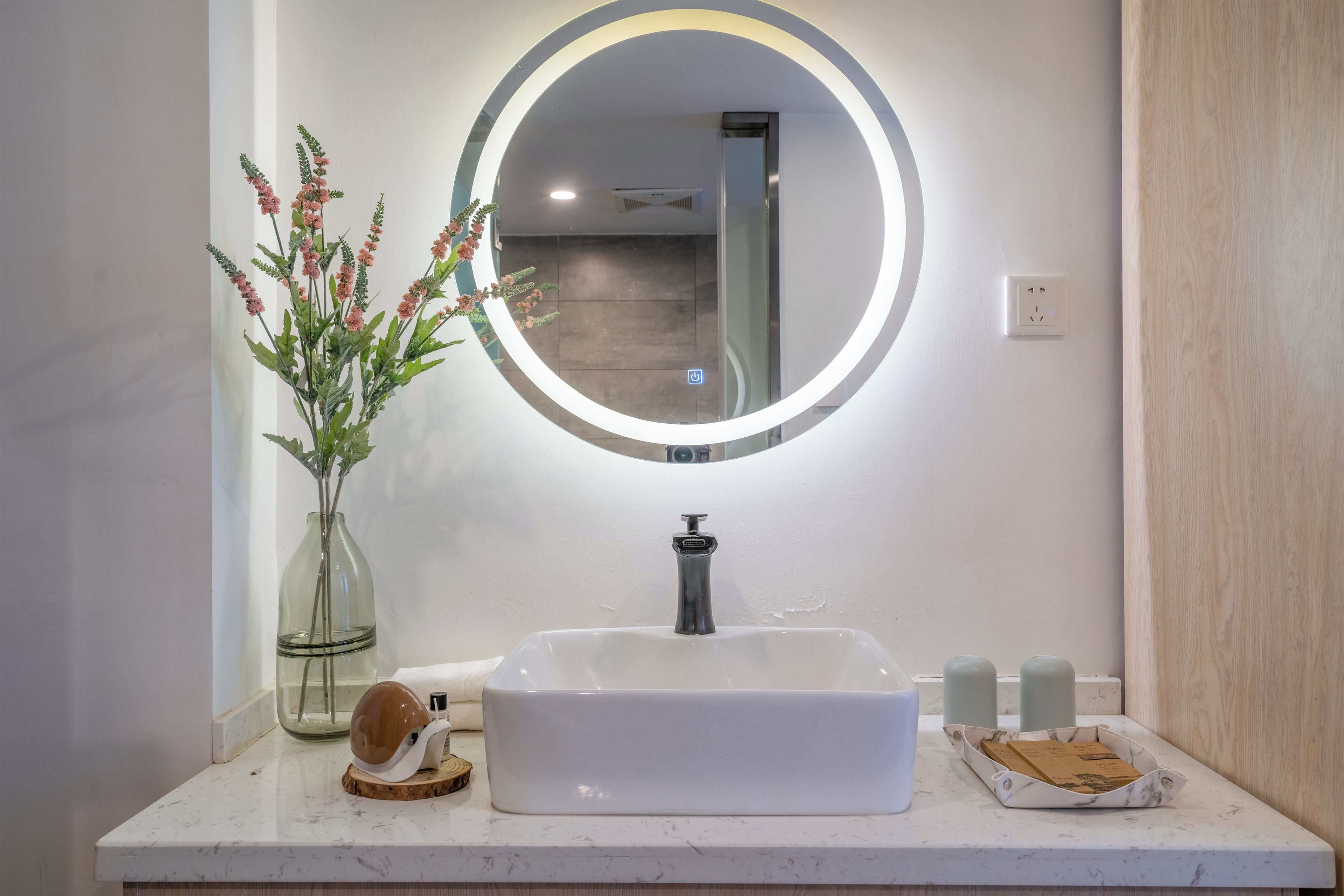 Un baño con un lavamanos blanco, un espejo redondo con luz, un florero con flores y un jabonera.