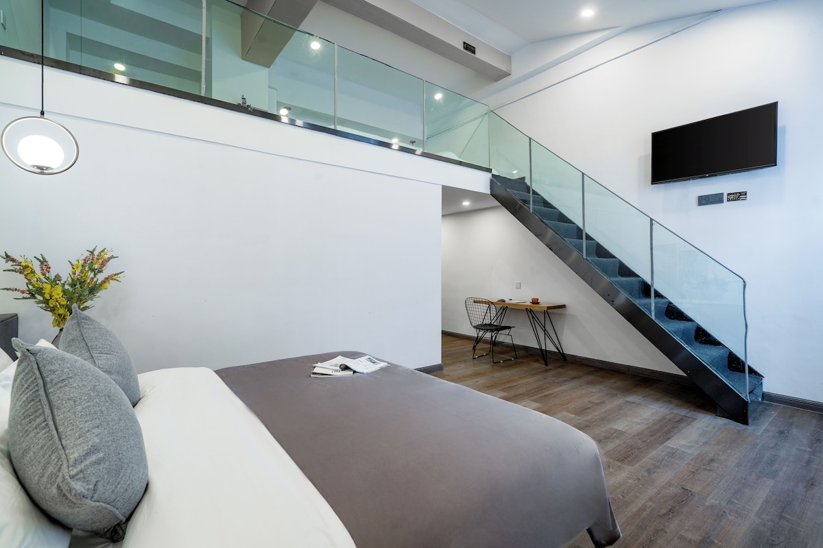 Een moderne slaapkamer met een bed, een klein tafeltje en een stoel. Een trap met glazen leuning loopt naar een hoger gelegen niveau.