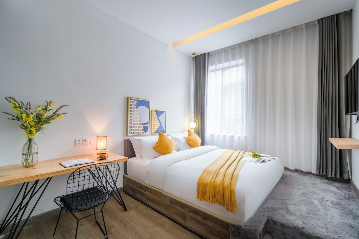 Een hotelkamer met een bed, een bureau, een stoel, een lamp, een vaas met bloemen en een schilderij aan de muur.