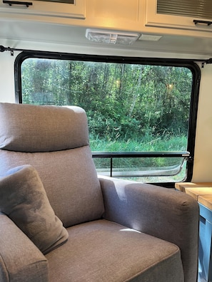 A gray cushioned seat with a window view of a forested area. - Serene 1-bedroom recreational vehicle on private property in Castle Rock (Castle Rock)
