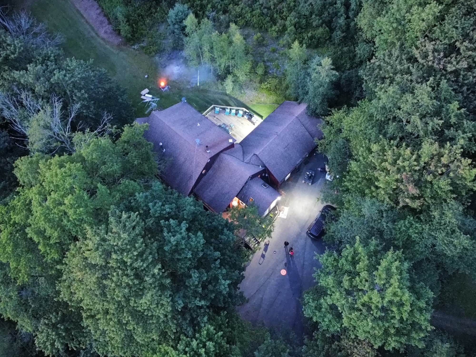 An aerial view of a house surrounded by dense forest, with a visible driveway and a small parking area.