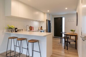 Uma cozinha moderna com bancada branca, três banquetas, uma mesa de jantar com duas cadeiras e uma porta preta ao fundo.