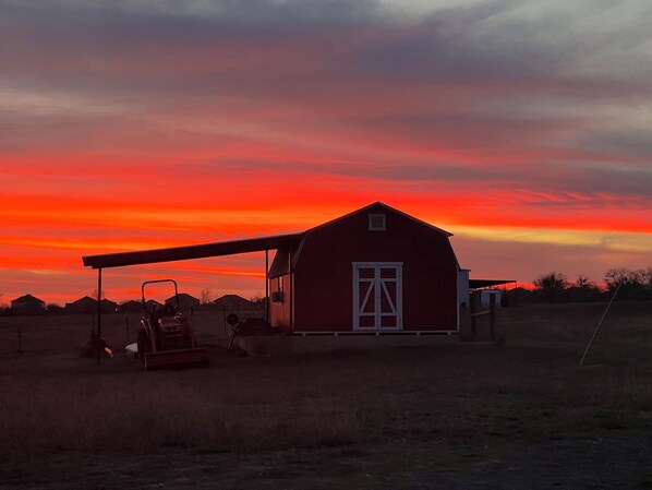 Un granero rojo con una puerta blanca, un tractor y un campo al atardecer.