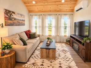Una sala de estar con sofá, mesa de centro, televisor y un ventanal con cortinas.