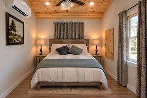 Un dormitorio con techo de madera, una cama con colcha estampada, dos lámparas de noche, una ventana con cortinas y un aire acondicionado instalado en la pared.
