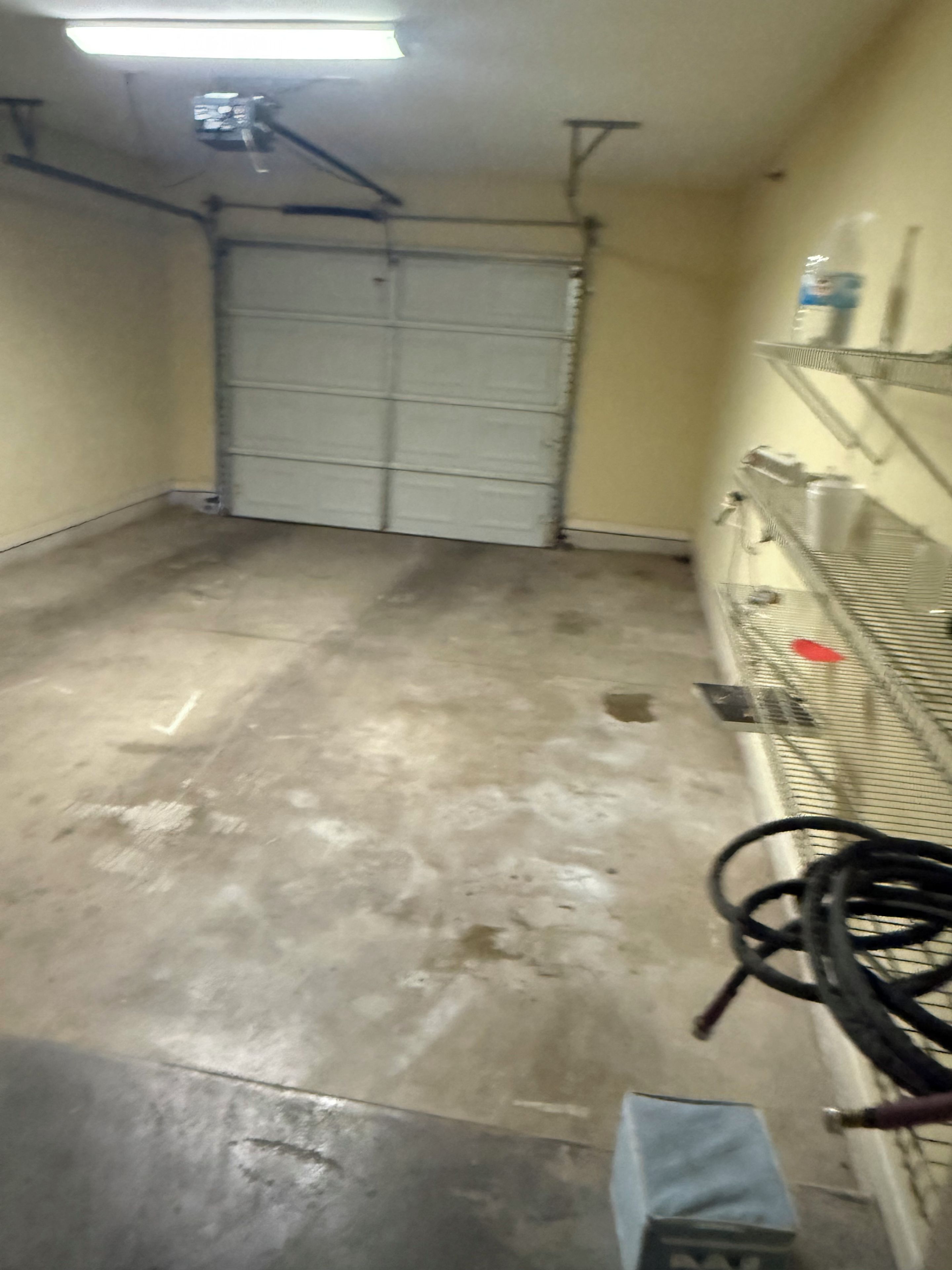 Un garage con pavimento in cemento, porta garage chiusa e una scaffalatura metallica con diversi oggetti.