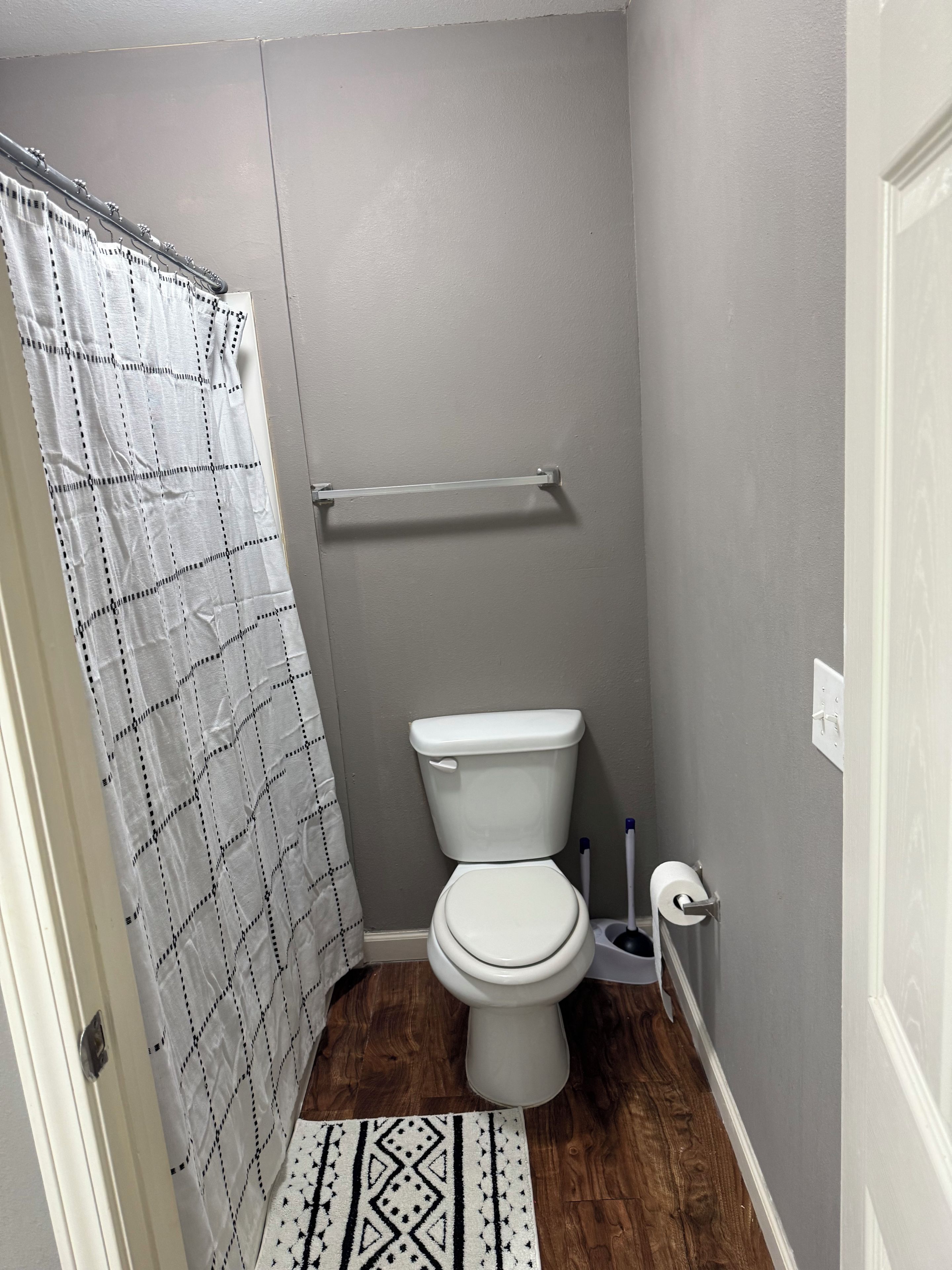 A small bathroom with a toilet, a shower curtain, a handrail, and a patterned rug.