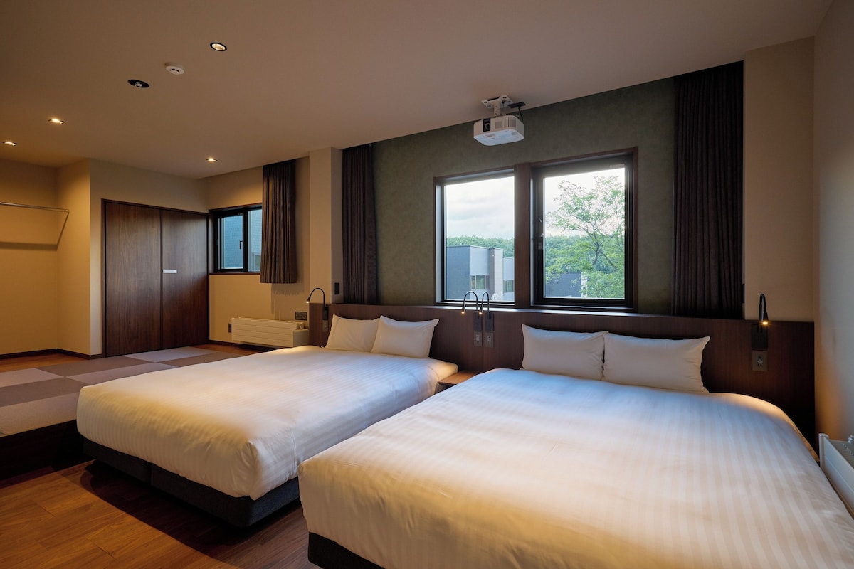 A hotel room with two beds, a large window, a projector, and a wardrobe.