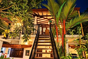 Una escalera conduce a un edificio con un nivel superior cubierto, rodeado de abundante vegetación.
