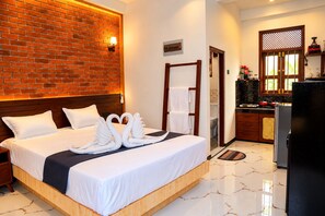 Habitación de hotel con pared de ladrillo, cama con ropa blanca, cabecera de madera, toallero, baño pequeño y kitchenette.