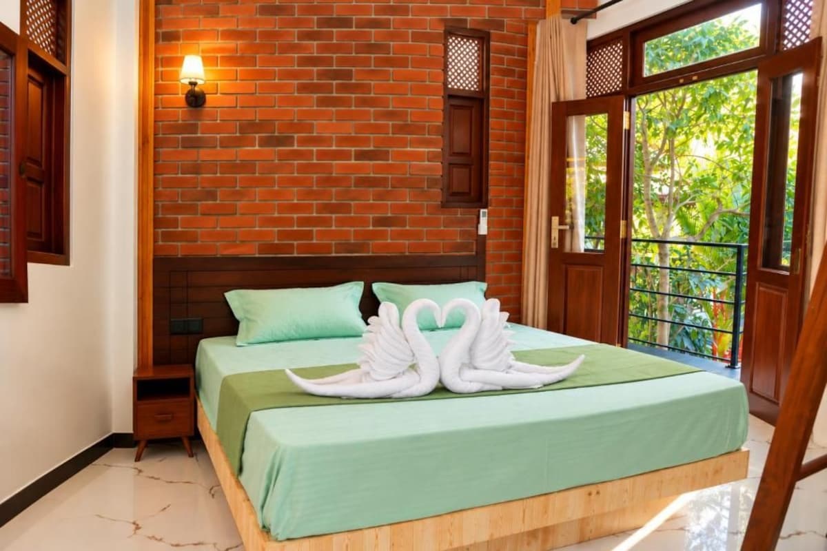 Habitación de hotel con pared de ladrillo, cama con cubrecama verde, mesita de noche, lámpara fijada en la pared y ventana con vista a la vegetación.