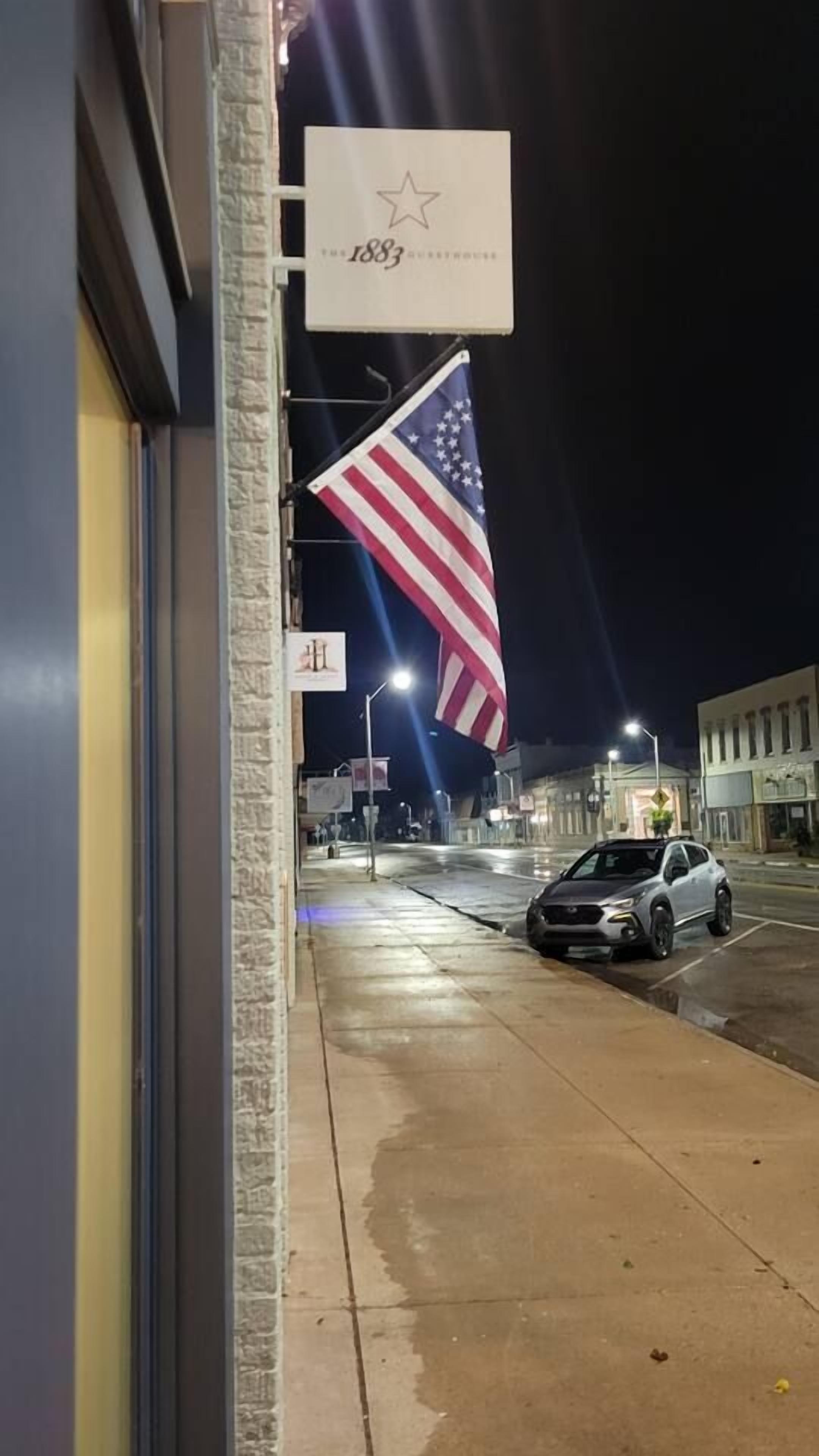 Vista de la calle por la noche con un coche aparcado, una gran bandera estadounidense y un edificio con un cartel que indica el año 1883.