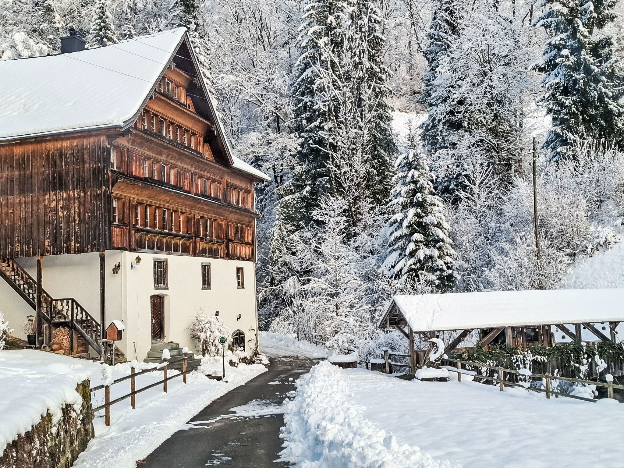 A traditional wooden house with a steeply pitched roof, a visible staircase, and a snow-covered path leading to a wooden bridge.