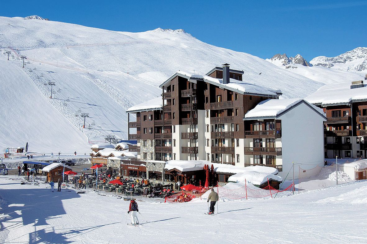 A snow-covered ski resort with multi-story buildings, outdoor seating areas, and a ski lift in the background.