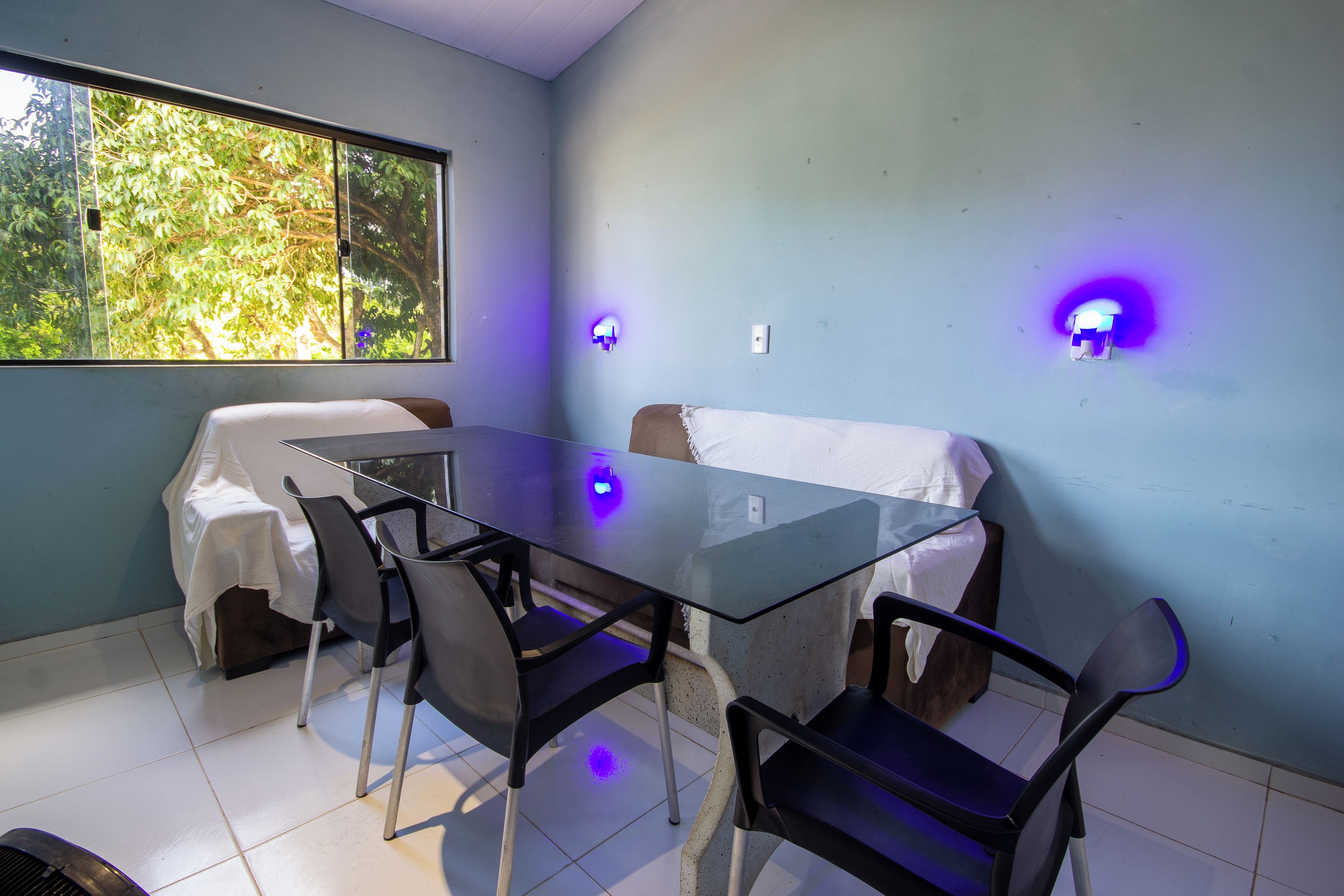A dining area with a glass table and chairs, a window with a view of trees, and wall-mounted lights.
