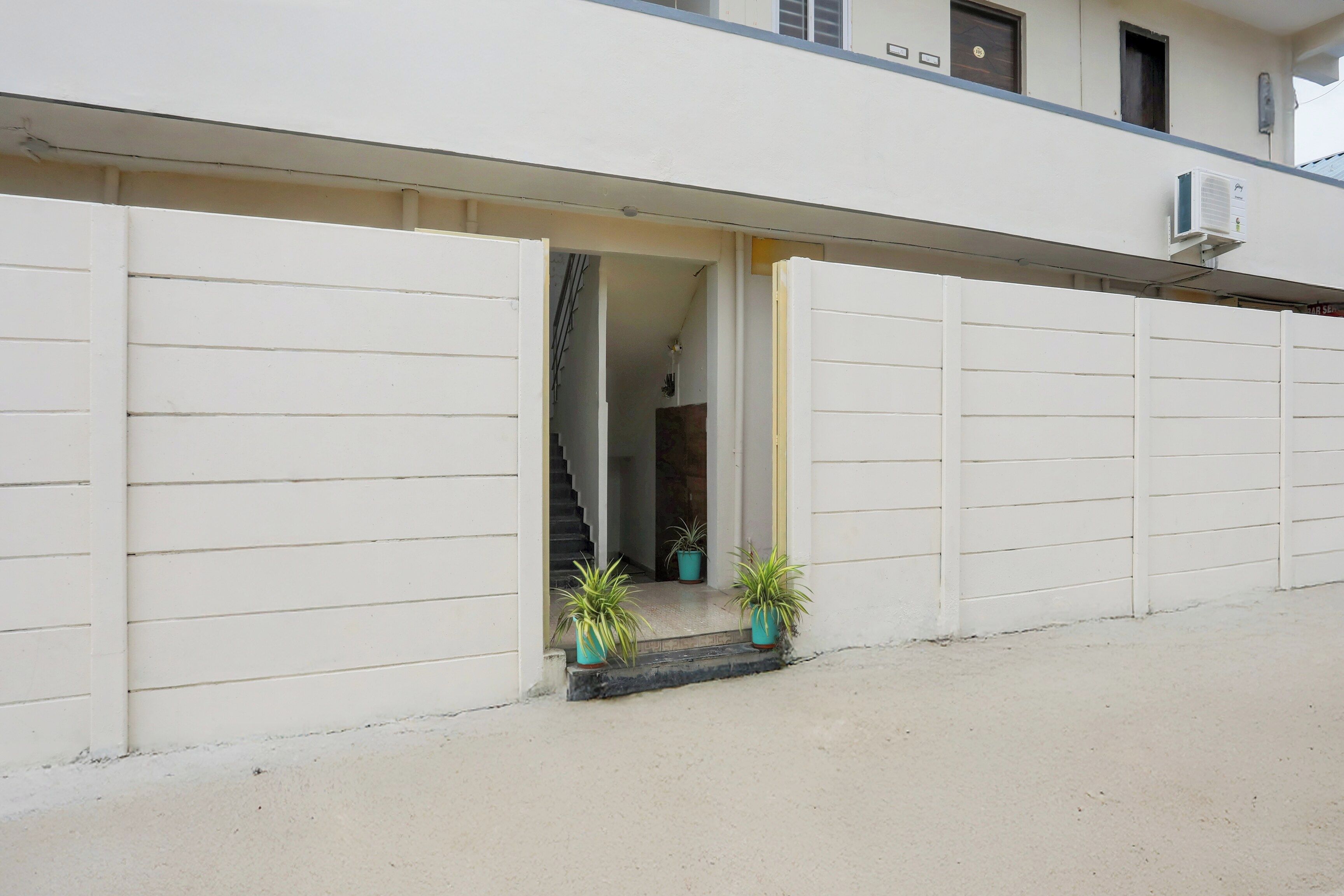 Un bâtiment à la façade blanche, avec un escalier apparent menant à un étage supérieur, et deux plantes en pot situées au rez-de-chaussée.