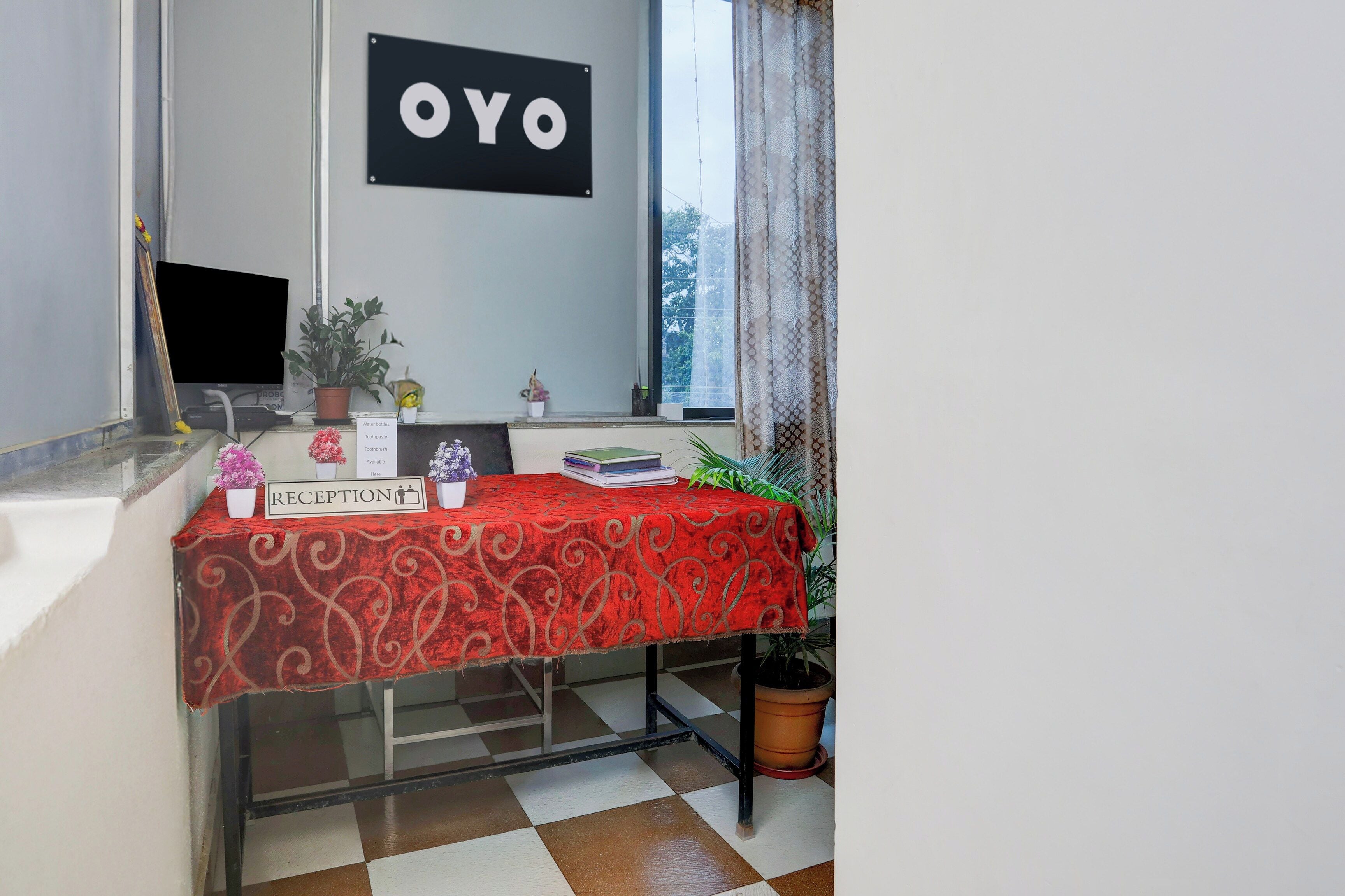 Uma área de recepção com uma mesa estampada em vermelho, um aviso indicando que garrafas de água e creme dental estão disponíveis, e um letreiro com 'OYO' na parede.