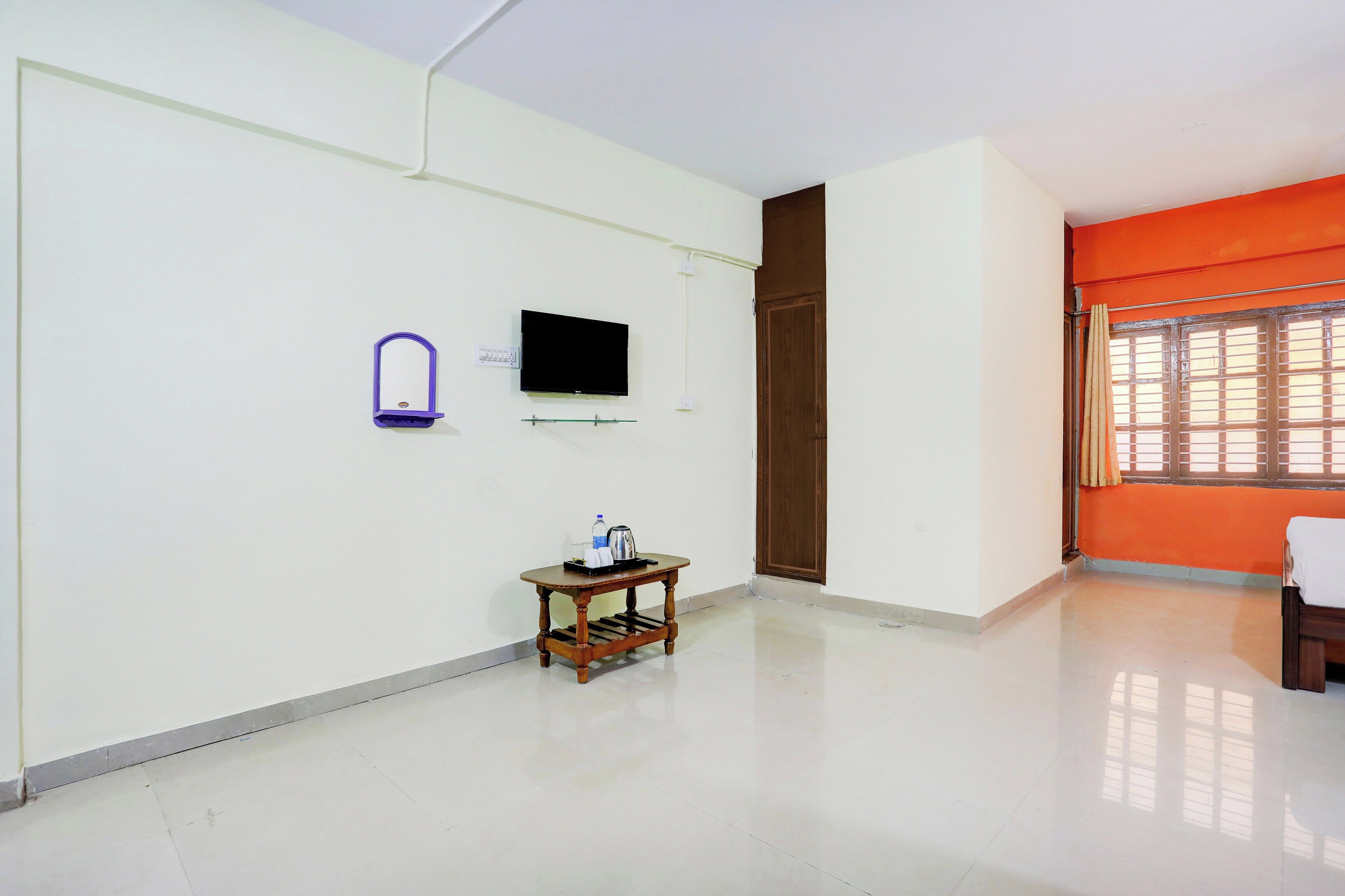 A room with a flat-screen TV mounted on the wall, a small wooden table with a water bottle and a kettle, and a door with a brown frame.