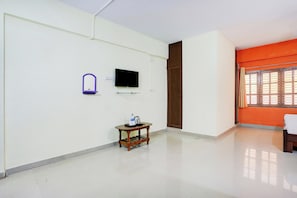 A room with a flat-screen TV mounted on the wall, a small wooden table with a water bottle and a kettle, and a door with a brown frame.