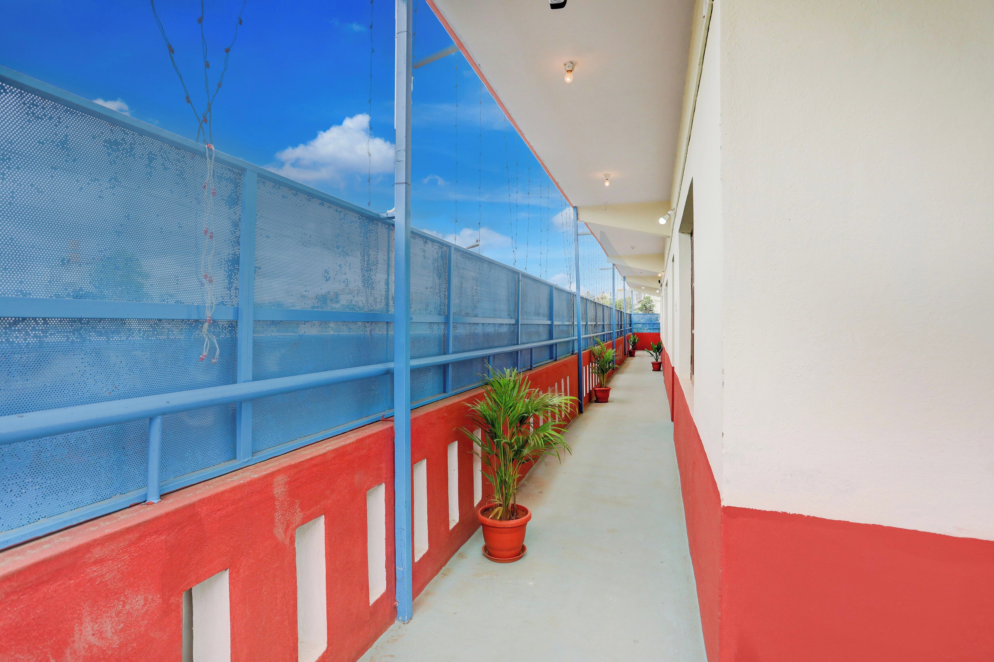A covered outdoor corridor with a blue mesh railing, potted plants, and a red and white wall.