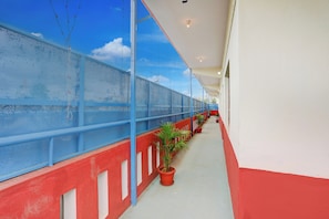 A covered outdoor corridor with a blue mesh railing, potted plants, and a red and white wall.