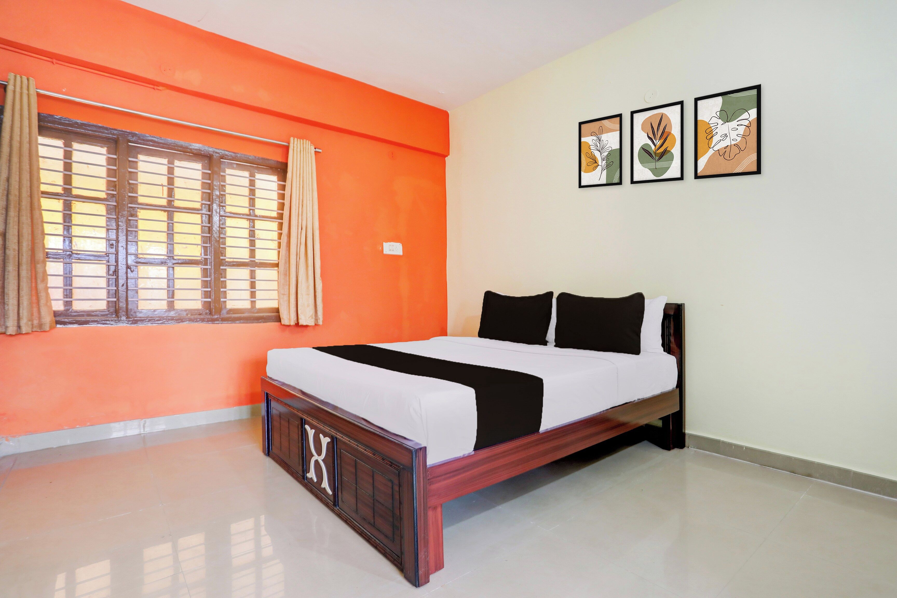 Un dormitorio con una pared naranja, una cama con ropa de cama blanca y negra, una ventana con cortinas y tres cuadros enmarcados en la pared.