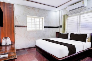 Habitación de hotel con una cama, una ventana con persianas, un equipo de aire acondicionado y un escritorio de madera con un juego de botellas de agua.