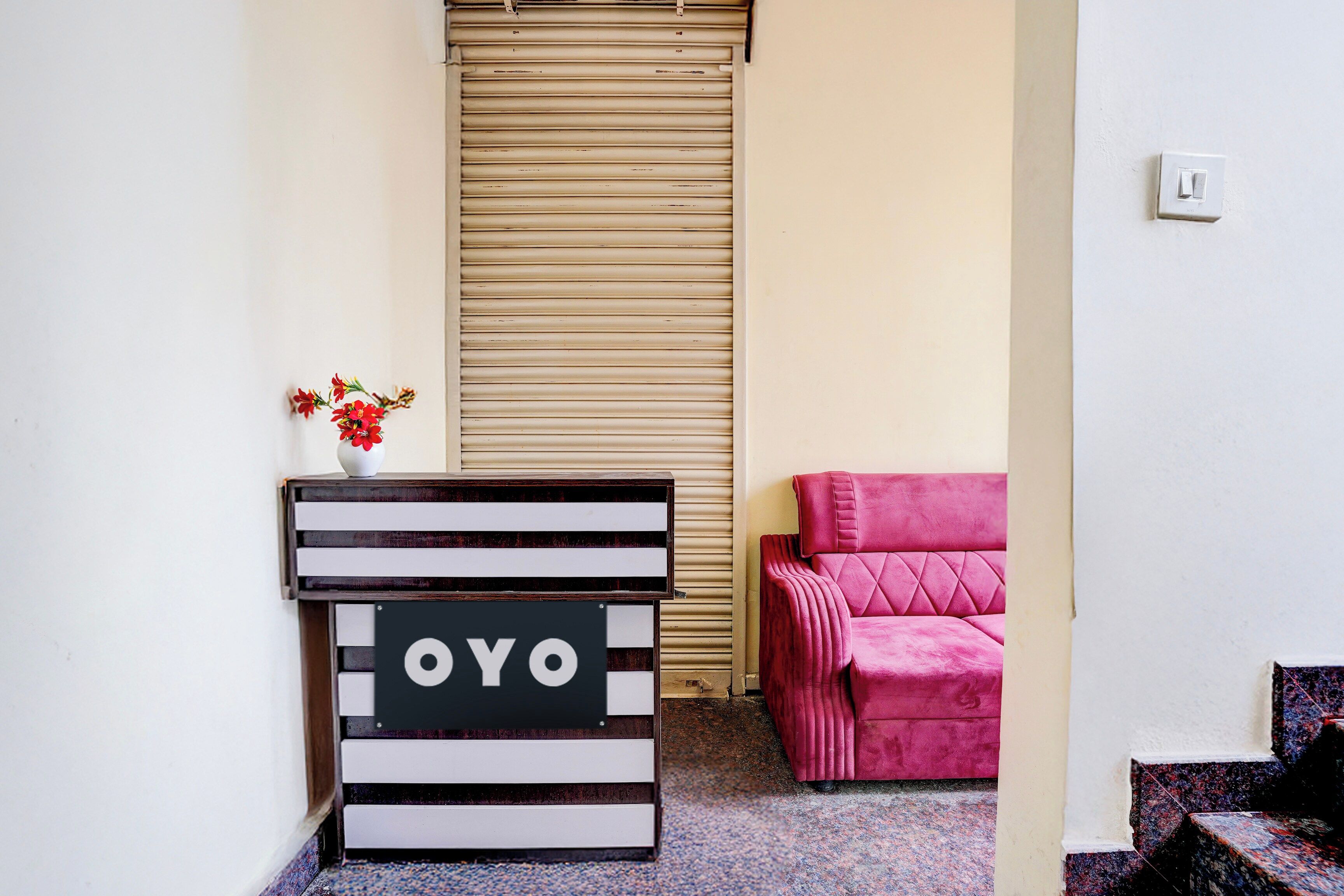 Une pièce avec une porte de volet fermée, une petite table sur laquelle est posé un panneau, une chaise rose et un escalier.