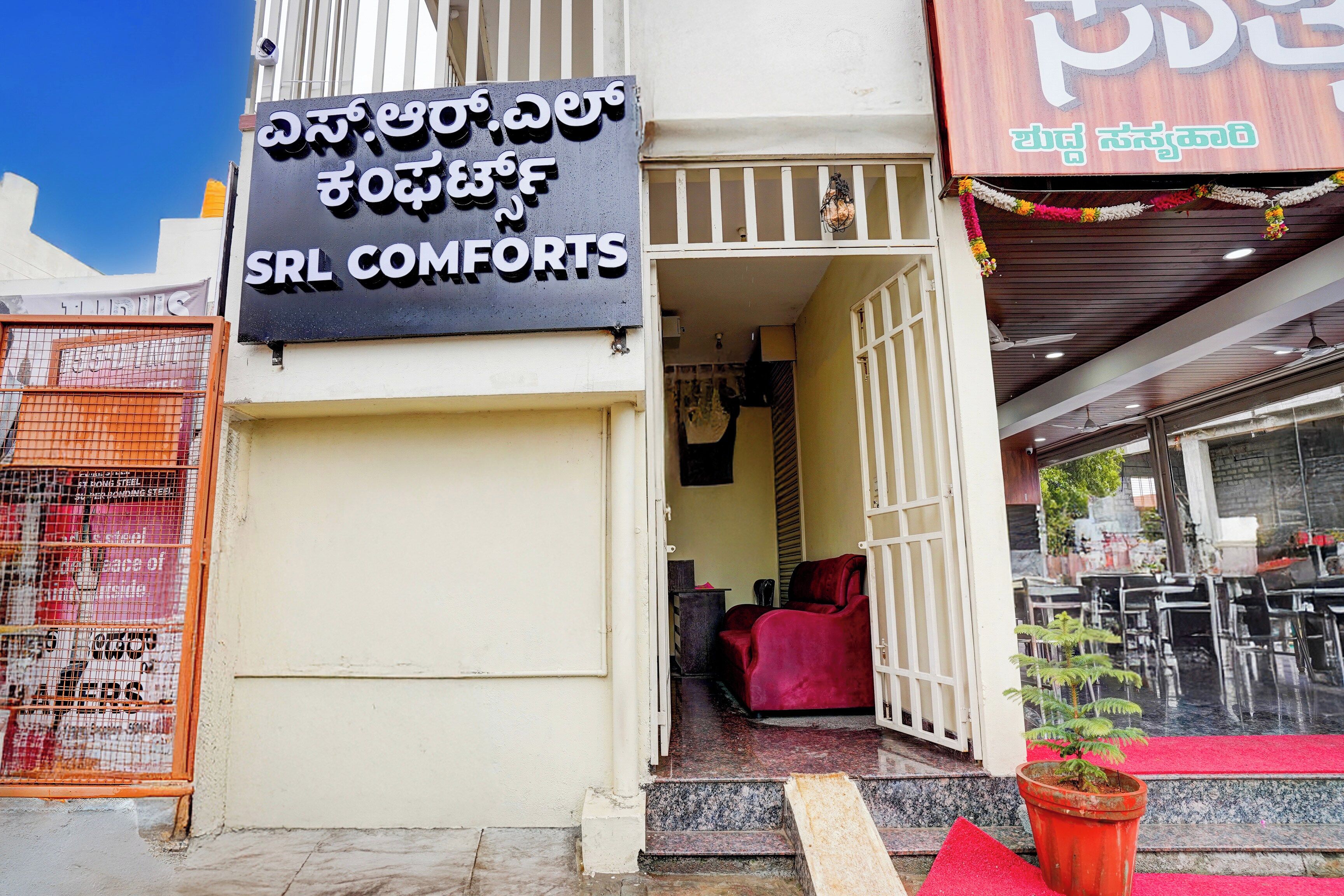 L’entrée d’un hôtel, avec une enseigne indiquant « Srl Comforts », une chaise rouge à l’extérieur et une petite plante en pot.