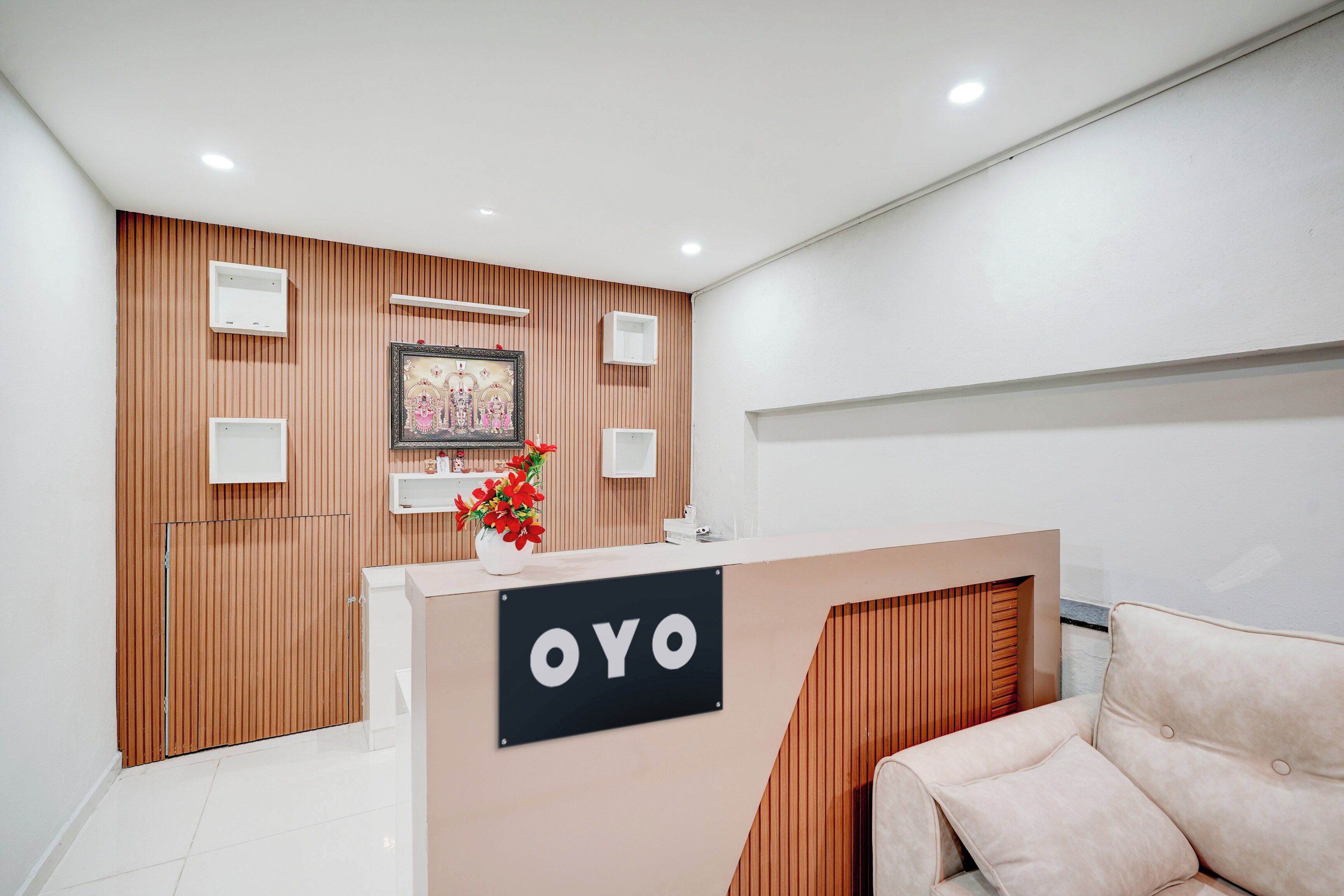 A reception area with a wooden panel wall, a reception desk with 'OYO' signage, a framed picture above the desk, and a potted plant with red flowers.