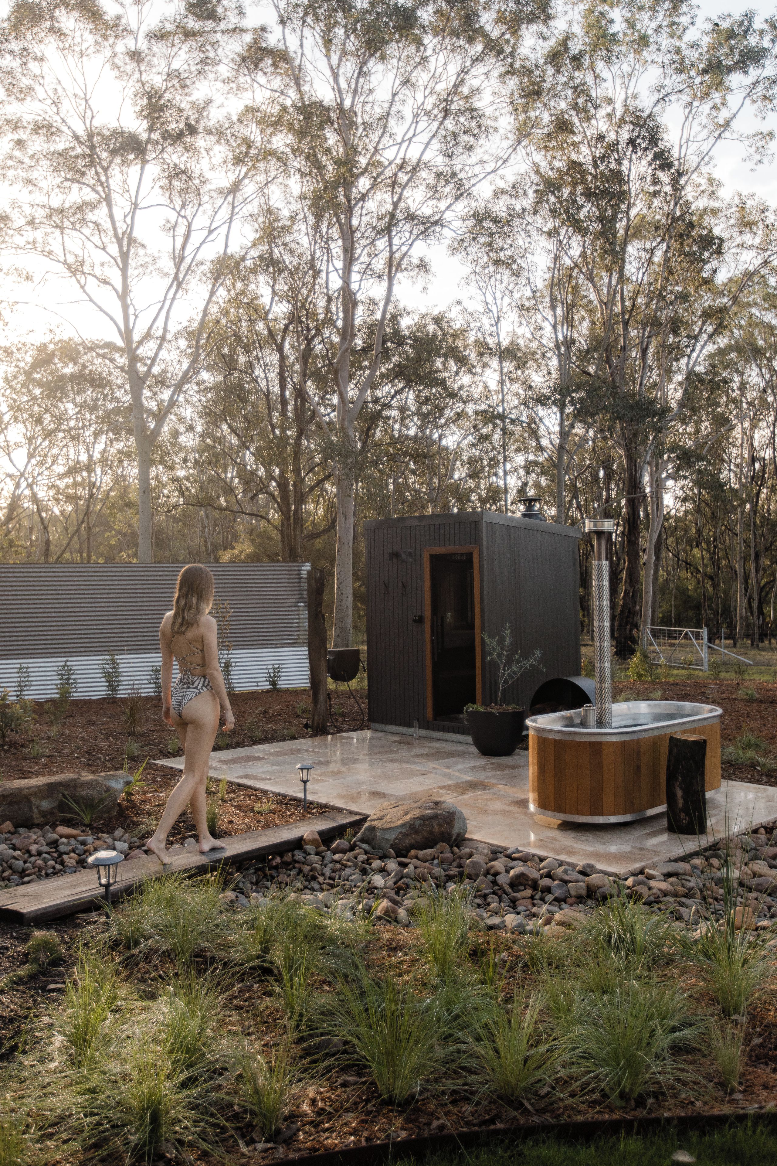 Una persona caminando sobre una terraza de madera con una sauna y una pequeña construcción de madera en un jardín.