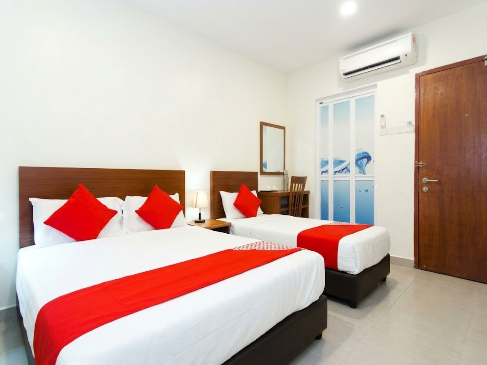 A hotel room with two beds, each with red and white bedding, a wooden headboard, a nightstand, a mirror, and an air conditioning unit.