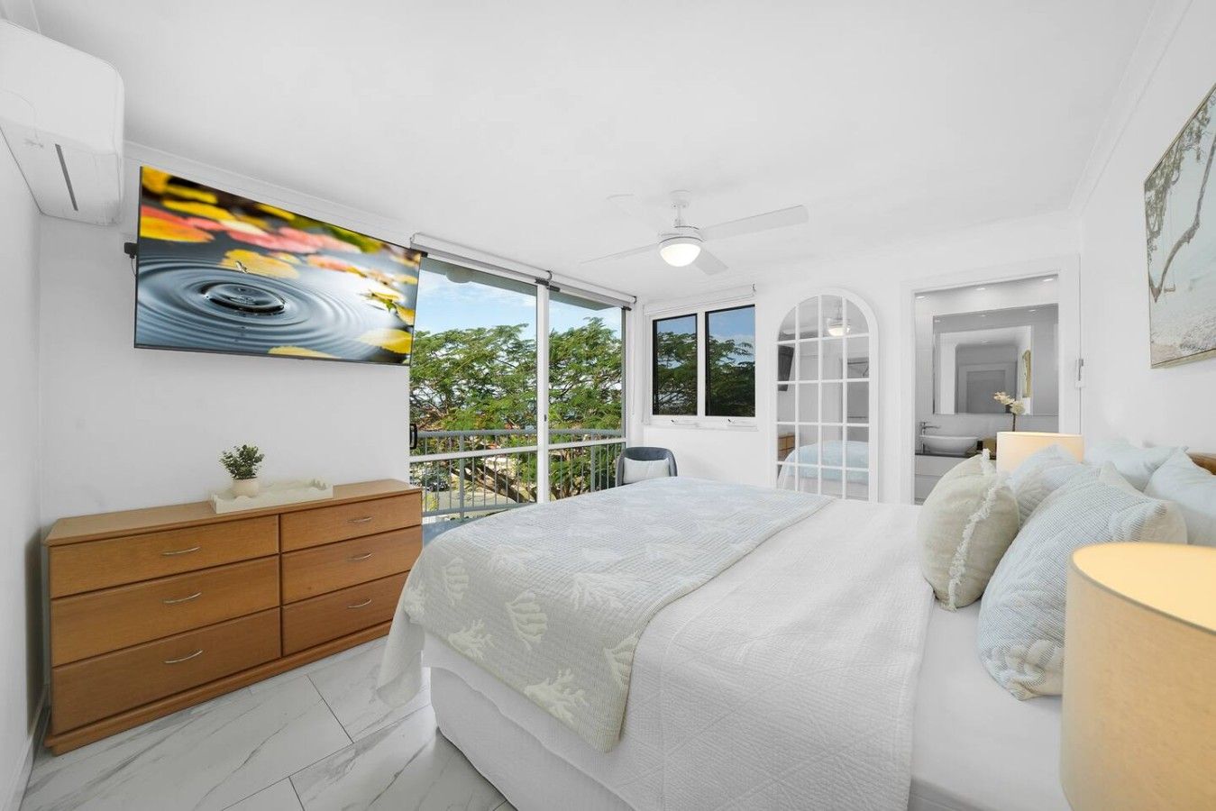 Een slaapkamer met een bed, een kast, een televisie en een balkon met uitzicht op bomen.