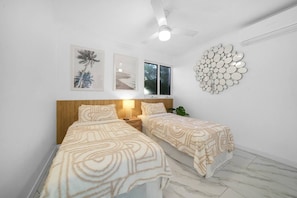 Un dormitorio con dos camas, un ventilador de techo, obras de arte colgadas en la pared y un espejo decorativo circular.