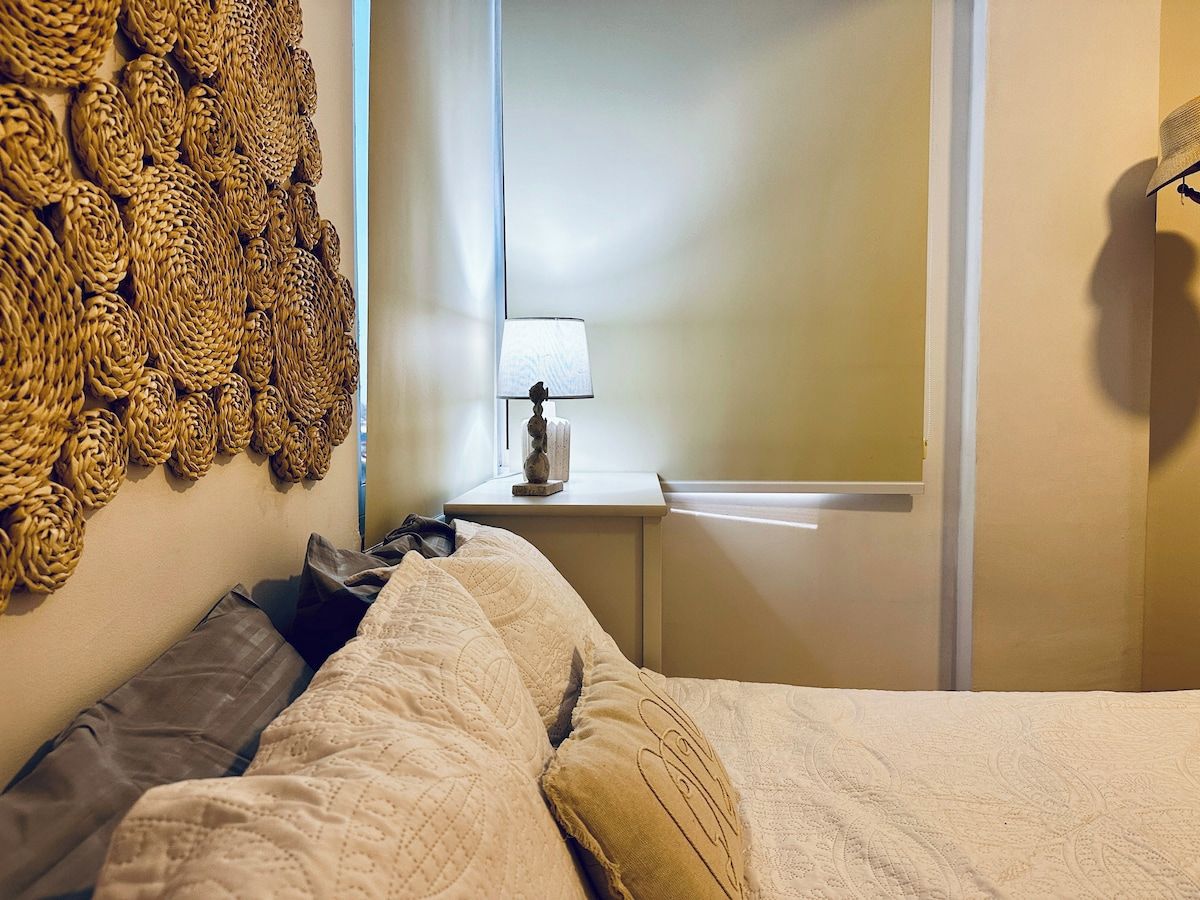 A bedroom with a bed, pillows, a nightstand with a lamp, and a wall with woven decorations.